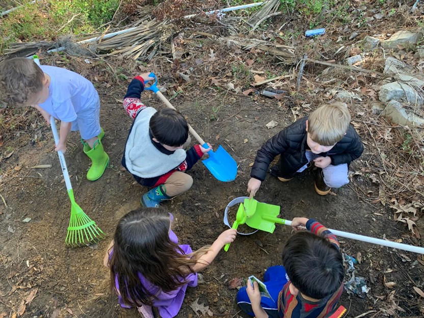 Five children with small shovels and rakes digging in a cleared area of dirt. 