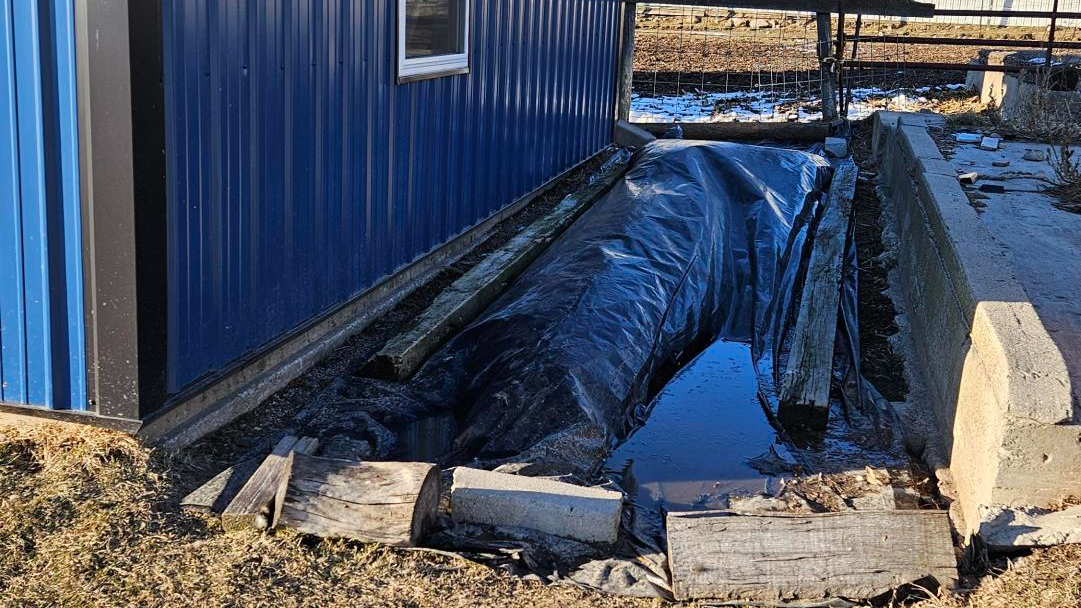 A garden site in the process of solarization, with a black plastic tarp spread across a trench to trap solar heat and eradicate weeds, pests, and soil pathogens. This preparation step is essential for creating a healthy garden bed. The site is adjacent to a blue metal-clad barn, and the area is bordered with old wooden beams and concrete blocks to secure the tarp in place. A small amount of snow in the background suggests this is being done in cooler seasons when the sun’s warmth is harnessed for soil treatment.